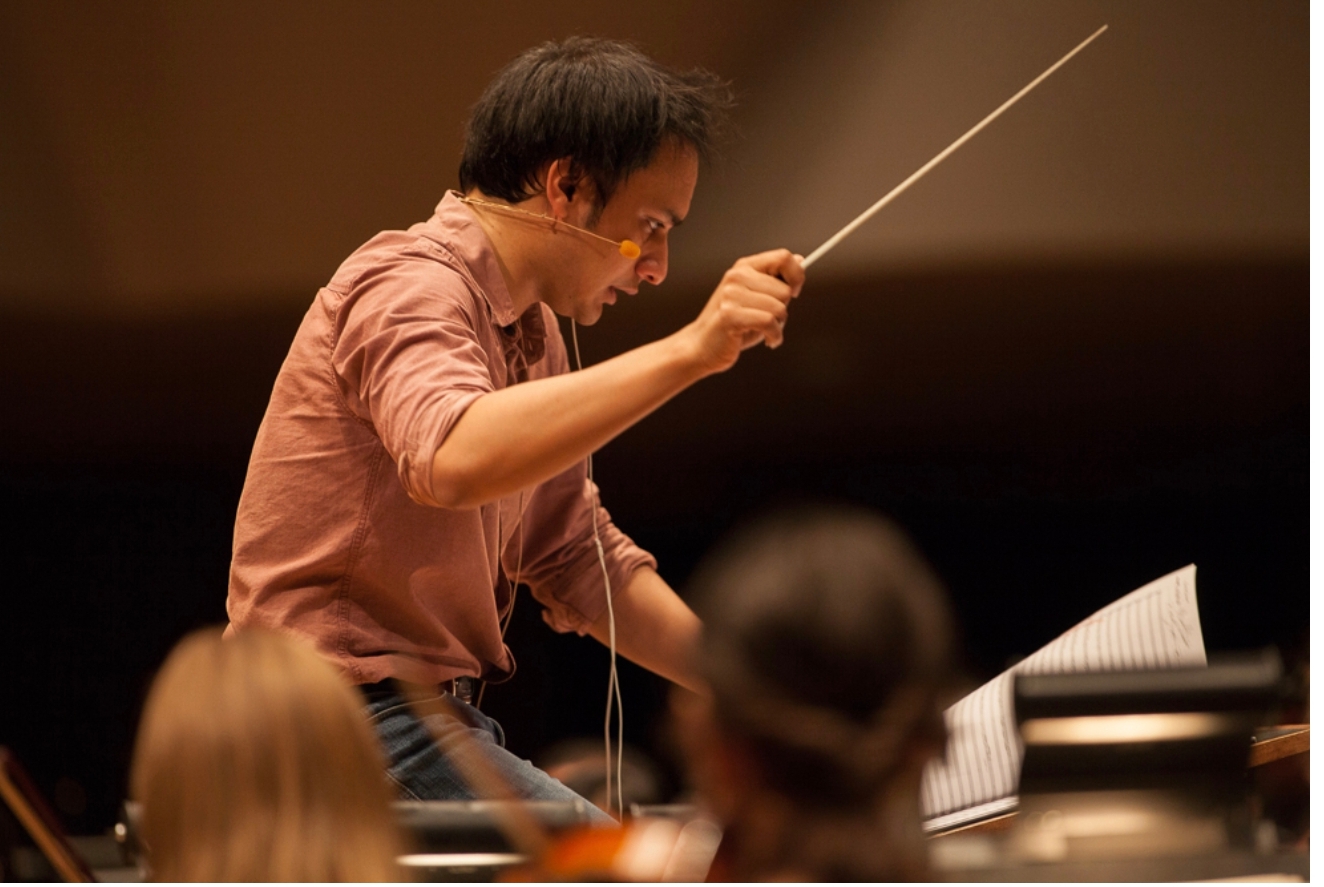 "O Maestro no Ensaio Geral": Fotografia de um maestro em plano médio curto, com gesto de grande exaustão/concentração, debruçado sobre a partitura e a batuta em ambiente de sala de concertos clássica.